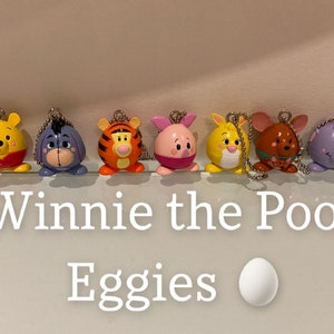 May include: A set of seven Winnie the Pooh character egg-shaped keychains. The characters include Winnie the Pooh, Piglet, Tigger, Eeyore, Rabbit, Owl, and Roo. The keychains are made of plastic and have a shiny finish. The text "Winnie the Pooh Eggies" is written below the keychains.