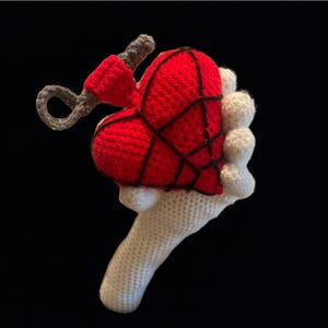 Puede incluir: Un juguete de peluche con forma de corazón tejido a crochet con un diseño en negro y rojo. El corazón está sostenido por una mano blanca tejida a crochet.