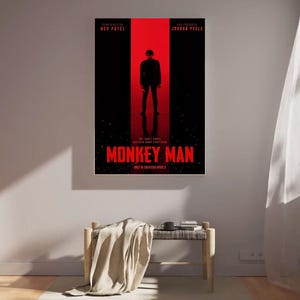 May include: A movie poster for "Monkey Man" featuring a silhouette of a person in a black suit against a red and black background. The title is in red, with the names Dev Patel and Jordan Peele above. The poster is displayed on a white wall.