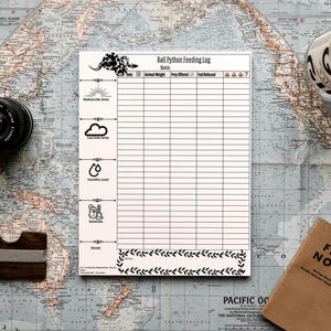 Op de afbeelding: Een Ball Python Feeding Log notitieblok met een decoratieve rand, een cameralens, een houten portemonnee, een Field Notes notitieboek en een fles op een vintage kaart. Het notitieblok heeft een slangenillustratie en secties voor het registreren van gegevens.