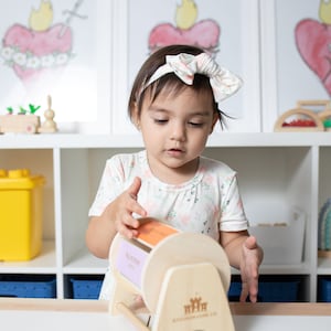 May include: A young child is playing with a wooden toy with colourful rotating panels. The toy has the words "My First Words" on it. The child is wearing a white shirt with a floral pattern and a white headband with a pink bow.