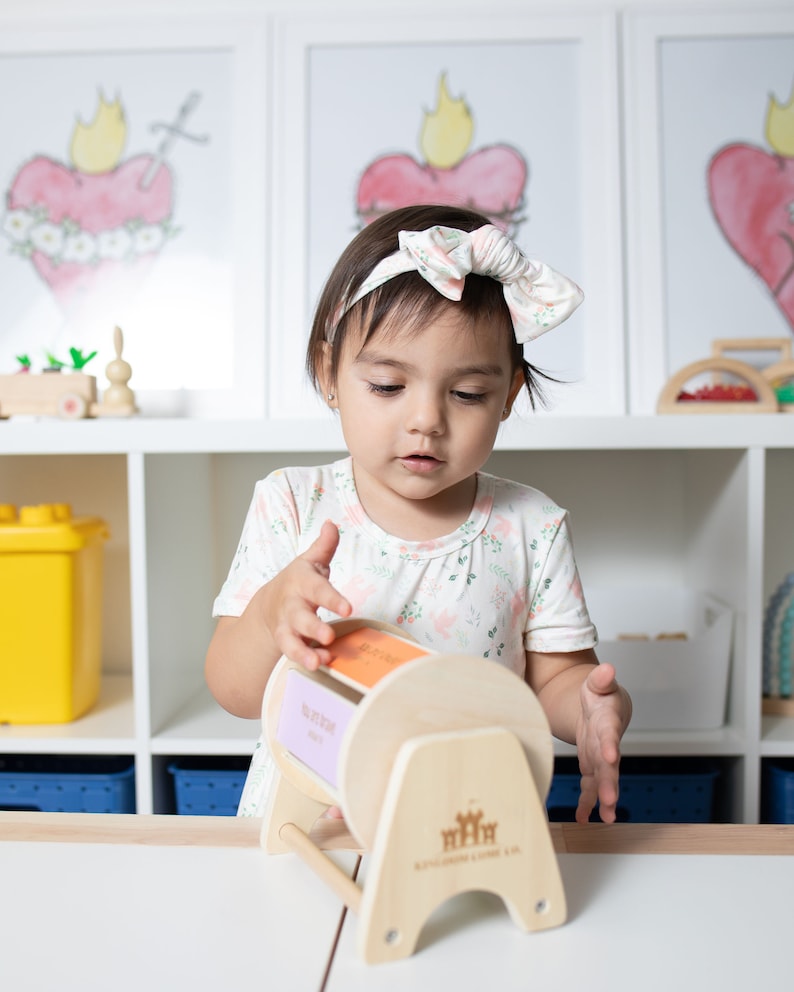May include: A young child is playing with a wooden toy with colourful rotating panels. The toy has the words "My First Words" on it. The child is wearing a white shirt with a floral pattern and a white headband with a pink bow.