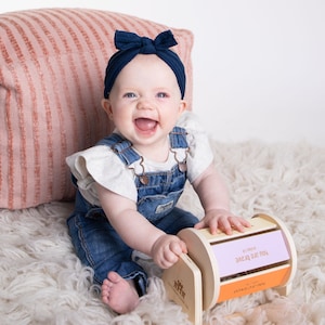 May include: A baby girl wearing a blue headband and denim dungarees is sitting on a white fluffy rug and playing with a wooden toy with the words "YOU ARE BRAVE" on it.