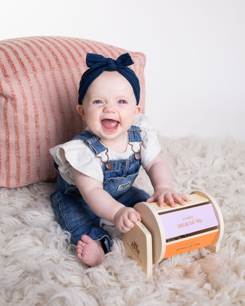 May include: A baby girl wearing a blue headband and denim dungarees is sitting on a white fluffy rug and playing with a wooden toy with the words "YOU ARE BRAVE" on it.