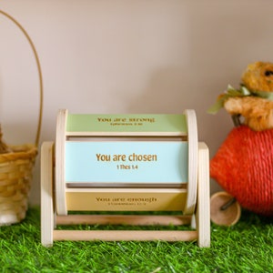 May include: A wooden spinning wheel with three panels, each with a different inspirational message. The top panel reads "You are strong", the middle panel reads "You are chosen 1 Thess 1:4", and the bottom panel reads "You are enough". The wheel is sitting on green grass with a basket and a stuffed bunny in the background.