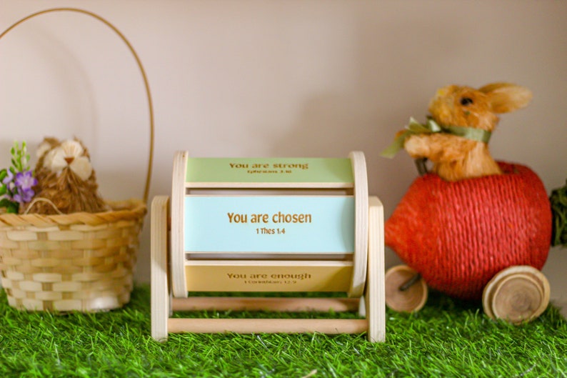 May include: A wooden spinning wheel with three panels, each with a different inspirational message. The top panel reads "You are strong", the middle panel reads "You are chosen 1 Thess 1:4", and the bottom panel reads "You are enough". The wheel is sitting on green grass with a basket and a stuffed bunny in the background.