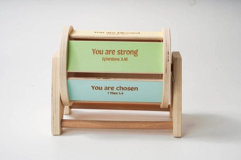May include: Wooden spinning wheel with three panels. The panels are painted in shades of green and blue. Each panel has a positive affirmation and a bible verse. The top panel reads "You are strong, Ephesians 3:16". The middle panel reads "You are blessed, Romans 12:2". The bottom panel reads "You are chosen, 1 Thes 1:4".