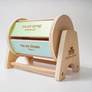 May include: Wooden spinning wheel with two panels, each featuring a different inspirational message and scripture reference. The top panel is light green and reads "You are strong" with the scripture reference "Ephesians 3:16". The bottom panel is light blue and reads "You are chosen" with the scripture reference "1 Thess 1:4". The wheel is mounted on a wooden stand with the words "Kingdom Come Co." and a castle logo.