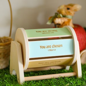 May include: A wooden toy with a rotating cylinder that displays three inspirational messages: "You are strong", "You are chosen", and "You are enough". The cylinder is painted in shades of green, blue, and yellow.