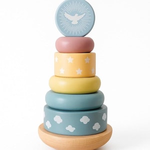 May include: A wooden stacking toy with a light blue top piece featuring a white dove design. The toy has various pastel-colored rings, including pink, yellow with white stars, and blue with white clouds. The base is a natural wood color.