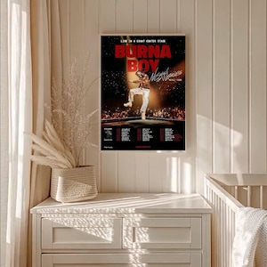 May include: A framed poster featuring Burna Boy on stage, with the text "BURNA BOY" in large red letters. The poster includes tour dates and the words "LIVE IN A GIANT CENTER STAGE". The image is displayed on a white wall.