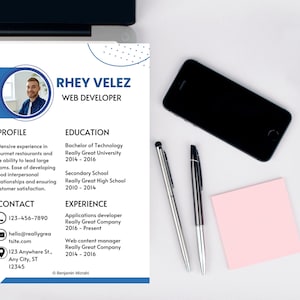 May include: A professional resume for Rhey Velez, a web developer, with a photo, contact information, and details about education and experience. The resume is on a white background with blue accents. A laptop, a smartphone, two pens, and a pink sticky note are also on the desk.