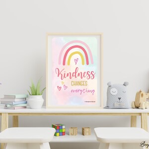 May include: A framed print with a rainbow design and the words "Kindness changes everything" in pink script. The artwork features a pastel colour palette and is displayed on a white table with decorative items.