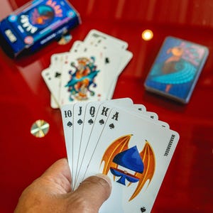 May include: A hand holding playing cards with a spade design, including the Ace, 10, Jack, Queen, and King of Spades. Two decks of cards are visible on a red surface. One deck is blue with a design, and the other has a colorful illustration.