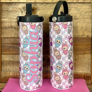 May include: Two stainless steel water bottles with black lids and handles. The bottles feature a repeating pattern of cartoon bunnies in various colors on a pink and white checkered background. The name "Sophia" is written in pink script.