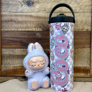 May include: A soft, light blue bunny figure with closed eyes and a smiling face sits beside a stainless steel water bottle. The bottle has a black handle and lid, and is decorated with a repeating pattern of bunny characters and the name "Coco" in pink.