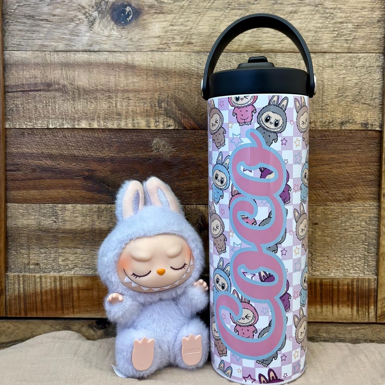 May include: A soft, light blue bunny figure with closed eyes and a smiling face sits beside a stainless steel water bottle. The bottle has a black handle and lid, and is decorated with a repeating pattern of bunny characters and the name "Coco" in pink.
