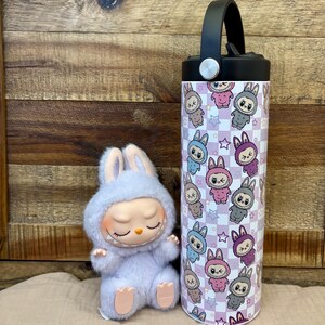 May include: A light purple bunny doll with closed eyes sits beside a stainless steel water bottle. The bottle has a black handle and a pattern of cartoon bunnies in various colours on a chequered background.