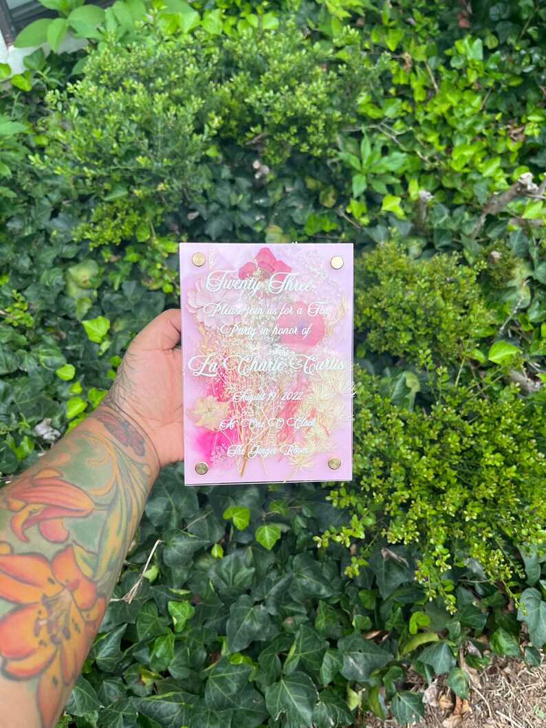 May include: A pink acrylic sign with a floral design and the text "Twenty Three", "Please join us for a Tea Party in honour of La' Charie Curtis", "August 19, 2022", "At One O'Clock", "The Ginger Room".