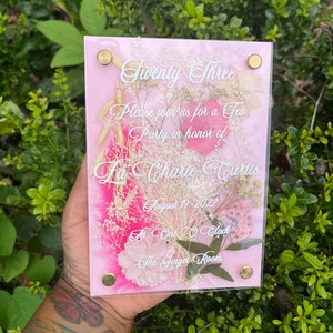 May include: A pink acrylic invitation with dried flowers pressed inside. The invitation reads "Twenty Three" and invites guests to a tea party in honour of La Charie Curtis on 19 August 2022 at 1:00 PM in The Ginger Room.