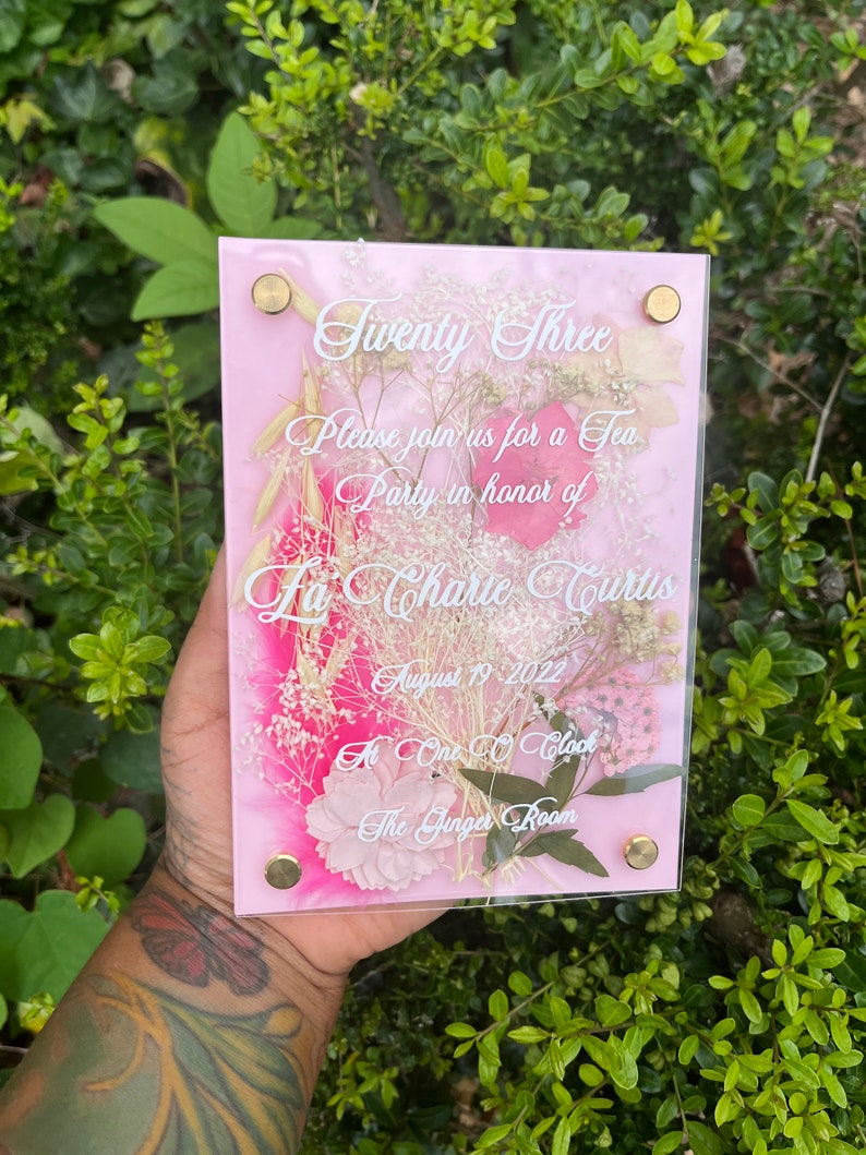 May include: A pink acrylic invitation with dried flowers pressed inside. The invitation reads "Twenty Three" and invites guests to a tea party in honour of La Charie Curtis on 19 August 2022 at 1:00 PM in The Ginger Room.
