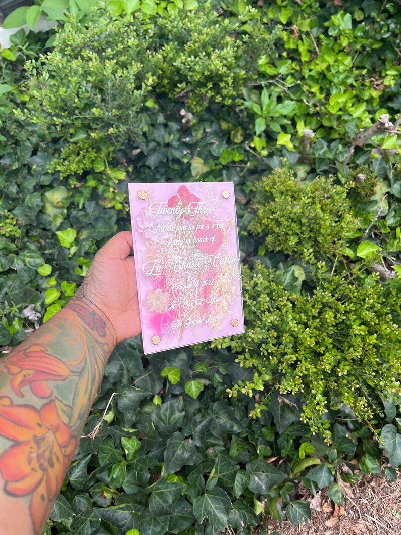 May include: A pink and gold acrylic invitation with a floral design and the text "Twenty Three" and "Please join us for a Tea Party in honour of La'Charte Curtis August 17, 2022 at 7:00 PM The Gage Room"