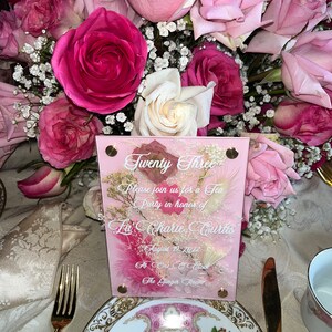 May include: A pink and white invitation card with the text "Twenty Three" and "Please join us for a Tea Party in honour of La Charie Curtis" on it. The date is August 19, 2022, and the time is One O'Clock. The location is The Ginger Room.