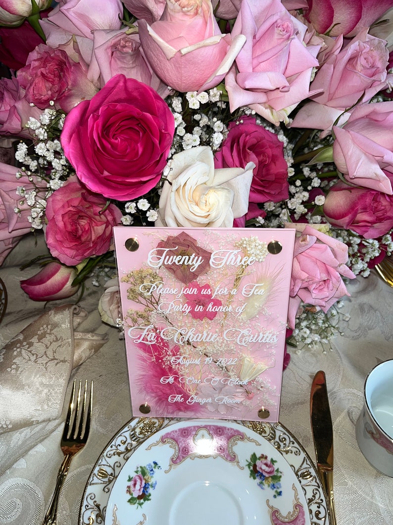 May include: A pink and white invitation card with the text "Twenty Three" and "Please join us for a Tea Party in honour of La Charie Curtis" on it. The date is August 19, 2022, and the time is One O'Clock. The location is The Ginger Room.