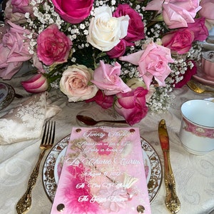 May include: A pink acrylic invitation with white text that reads "Twenty Three Please join us for a Tea Party in honour of La Charie Curtis August 9, 2022 At One O'Clock The Ginger Room". The invitation is decorated with dried flowers and glitter.