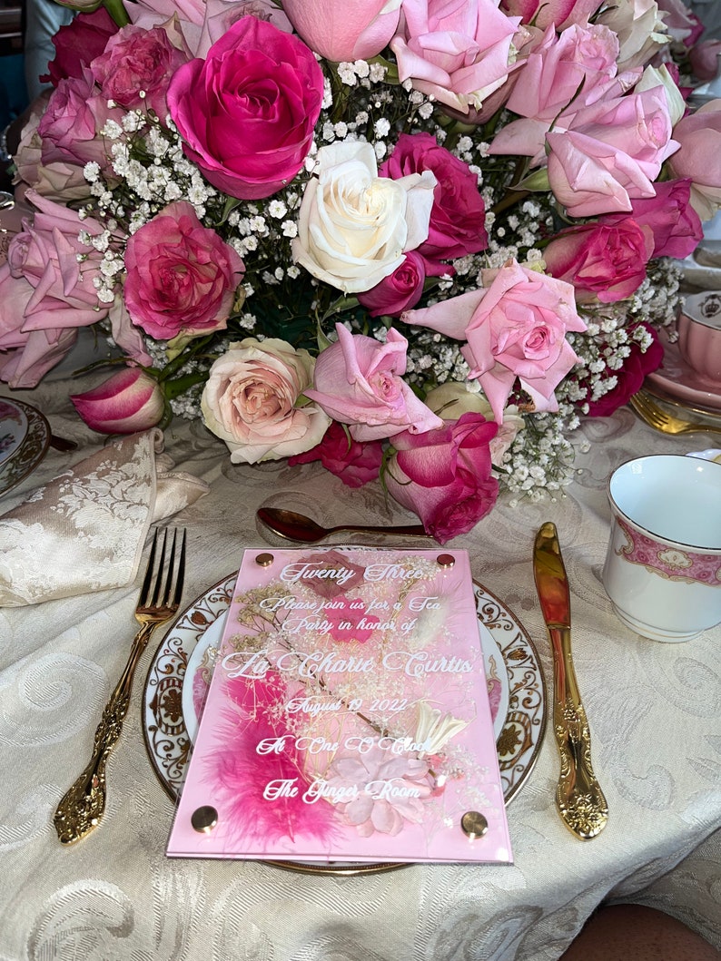 May include: A pink acrylic invitation with white text that reads "Twenty Three Please join us for a Tea Party in honour of La Charie Curtis August 9, 2022 At One O'Clock The Ginger Room". The invitation is decorated with dried flowers and glitter.