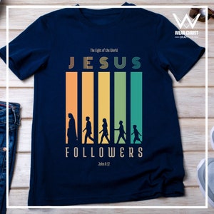 Puede incluir: Camiseta azul marino con un gráfico de estilo retro de una silueta de Jesús y sus seguidores. El gráfico incluye el texto "The Light of the World", "JESUS", "FOLLOWERS" y "John 8:12".