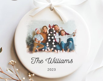 May include: White ceramic Christmas bauble featuring a photograph of a family and their dog, with the text "The Williams" and the year "2023".