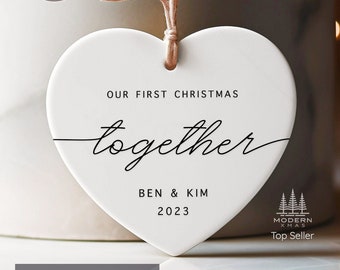Könnte beinhalten: Weißer herzförmiger Keramik-Anhänger mit dem Text "Unser erstes Weihnachten zusammen" und den Namen "Ben & Kim" sowie dem Jahr "2023". Der Anhänger hat eine braune Kordel zum Aufhängen. Der Text "Modern Xmas Top Seller" ist ebenfalls sichtbar.
