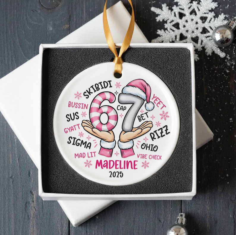 May include: A white ceramic Christmas ornament with a gold ribbon, presented in a gift box. The ornament features the number "67" in pink and gray, with text including "Madeline" and "2025". The design includes snowflakes and various slang terms.