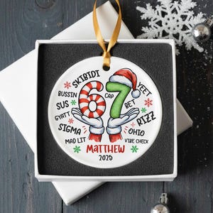 May include: A white ceramic Christmas ornament with a gold ribbon, presented in a gift box. The ornament features the number "67" in a candy cane design and green with a Santa hat. Text includes "SKIBIDI", "Matthew", and "2025".
