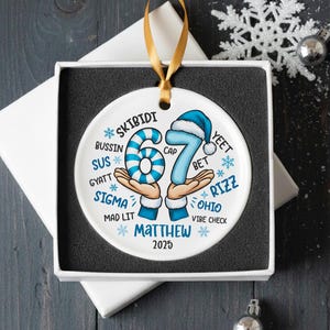 May include: A white ceramic ornament with a gold ribbon, presented in a gift box. The ornament features the number 67 in blue, with a blue and white striped 6 and a 7 wearing a blue cap. Text includes "SKIBIDI", "Matthew", and "2025".