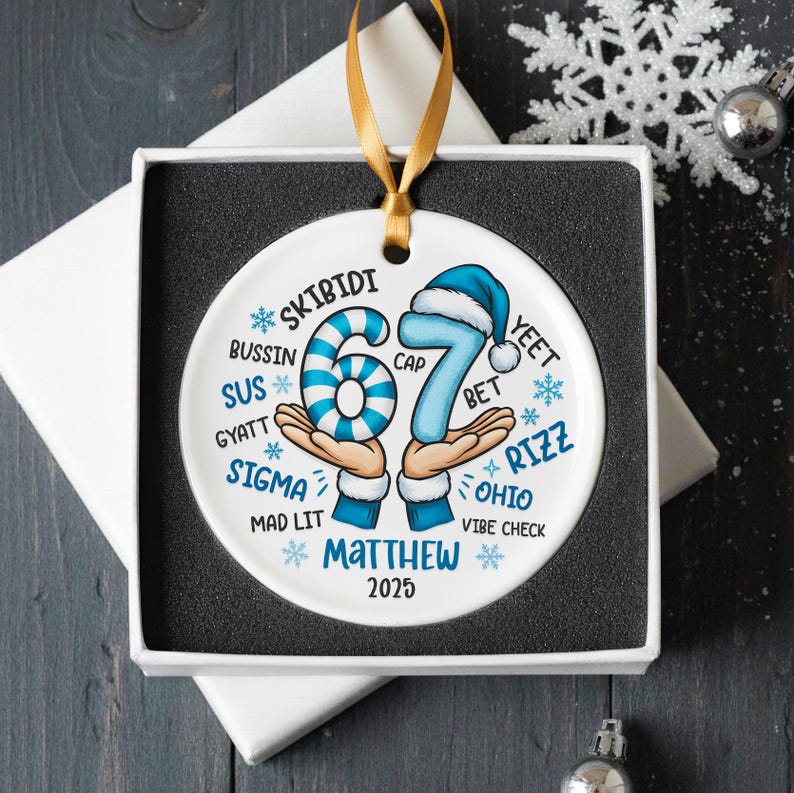 May include: A white ceramic ornament with a gold ribbon, presented in a gift box. The ornament features the number 67 in blue, with a blue and white striped 6 and a 7 wearing a blue cap. Text includes "SKIBIDI", "Matthew", and "2025".