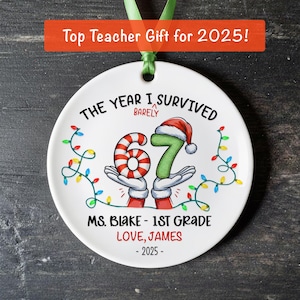 May include: A white ceramic ornament with a green ribbon for hanging. The ornament features the text "THE YEAR I SURVIVED BARELY" and the numbers "67" in a festive design, with the text "MS. BLAKE - 1ST GRADE LOVE, JAMES - 2025".