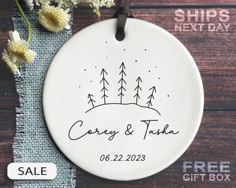 May include: A white ceramic ornament with a black ribbon for hanging. The ornament features a minimalist design of trees and stars, with the names "Corey & Tasha" and the date "06.22.2023" in script. The word "SALE" is visible.