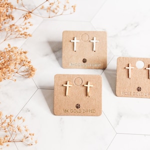 May include: Three pairs of gold-colored cross stud earrings displayed on individual brown cards. The cards are labeled with text indicating the type of gold used. The earrings are arranged on a white marble surface with dried floral accents.