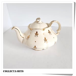 May include: Cream-colored ceramic teapot with a lid, spout, and handle, adorned with gold fleur-de-lis and dot patterns. The rim and handle are also trimmed in gold. The text "COLLECTA-BITS" is at the bottom.