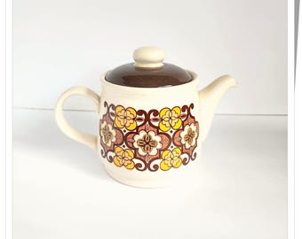 May include: A white ceramic teapot with a brown lid and a floral pattern in shades of yellow, brown, and orange. The teapot has a rounded body and a spout on the side.