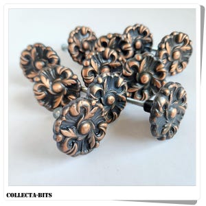 May include: A cluster of decorative cabinet knobs in a bronze finish. Each knob has an intricate floral pattern and a dark, aged appearance, attached to a silver screw. The knobs are displayed on a white background. The text "COLLECTA-BITS" is visible.