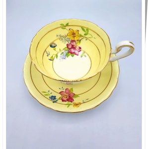 May include: A yellow teacup and saucer with a floral pattern. The cup and saucer have a gold trim and a scalloped edge.