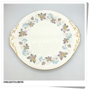 Può includere: Un piatto da torta in ceramica bianca con un bordo dorato e un motivo floreale con foglie blu, rosa e grigie. Il piatto ha due manici.