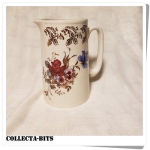 May include: A white ceramic pitcher with a floral design featuring red, pink, and blue flowers. The pitcher has a gold trim around the top and a handle.