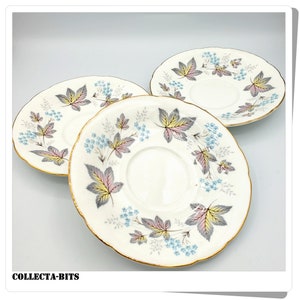 Può includere: Tre piattini in ceramica bianca con bordo dorato e un motivo floreale di foglie e fiori grigi, rosa e blu.