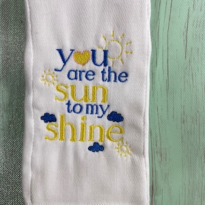 Può includere: Un bavaglino bianco con ricamo blu e giallo che recita "You are the sun to my shine" con un cuore, un sole e delle nuvole.