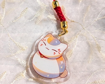以下が含まれることがあります： 赤い首輪と金色のアクセントが付いた、漫画の猫が特徴の透明なアクリルチャーム。猫は白い体で、オレンジ、青、金色のディテールがあります。金色の金属製ハードウェアが付いた赤いコードから吊り下げられています。