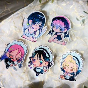 Könnte beinhalten: Eine Sammlung von sechs Aufklebern im Anime-Stil. Die Aufkleber zeigen verschiedene Charaktere mit Flügeln, Hörnern und Maid-Outfits. Die Farbpalette umfasst Blau-, Rosa- und Schwarztöne. Die Aufkleber befinden sich auf einer weiß-goldenen Oberfläche.